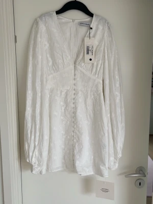 Vit blommig klänning från Adoore - Supersöt vit klänning från Adoore med blommigt broderat mönster, v-ringning och dekorativa knappar framtill. Klänningen har långa puffärmar och är tillverkad i ett lätt tyg som känns lyxigt. Perfekt för sommarens alla tillfällen SÄRSKILLT STUDENTEN🥳🎓!  Aldrig använd, helt ny med ettiketter 