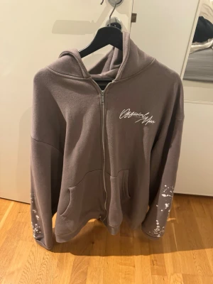 Brun zip-hoodie med tryck - Hoodien har vit text på bröstet och huvan, samt coola vita detaljer på ärmarna. Utrustad med huva och två fickor framtill. Perfekt för en avslappnad streetstyle-look.