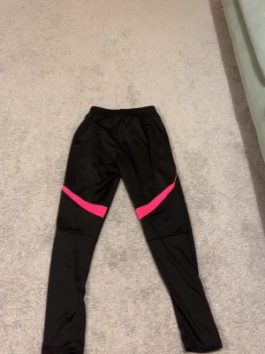 Svarta träningsbyxor med rosa detaljer - Snygga svarta träningsbyxor med rosa detaljer och dragkedja vid benslut. Byxorna har elastisk midja och tre rosa ränder på ena benet, samt rosa band över knäna. Perfekta för gymmet eller löpning. Passar både M och S men helst S!