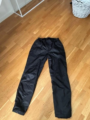 Svarta täckbyxor från Ski Industries - Svarta täckbyxor från Ski Industries med elastisk midja och raka ben. Byxorna är fodrade och tillverkade i ett vind- och vattenavvisande material, perfekta för kalla dagar. Klassisk design med fickor och tryckknapp i midjan.