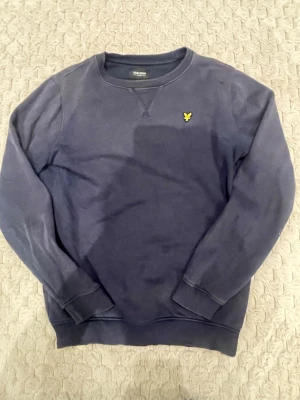 Mörkblå sweatshirt från Lyle & Scott - Klassisk mörkblå sweatshirt från Lyle & Scott med rund halsringning och gul logga broderad på bröstet. Tröjan har ribbade muddar vid ärmslut och nederkant, perfekt för en avslappnad och stilren look. Tröjan är också lite solblekt.