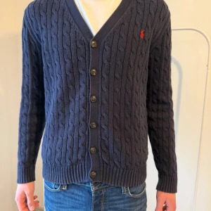 Mörkblå kabelstickad kofta Polo Ralph Lauren - Kabelstickad mörkblå kofta från Polo Ralph Lauren med v-ringning och knäppning framtill. Köpt på Åhléns barnavdelning största storleken 18-20 som motsvarar xl för 1699kr! Vårt pris endast 799!✅.          Tröjan är sparsamt använd och inga deffekter!  Kan diskutera pris vid snabb affär✅Fråga vid minsta lilla svarar snabbt! // mvh Hugo’s Wardrobe