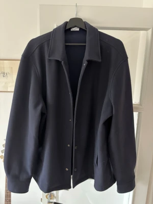 Mörkblå overshirt med tryckknappar - Stilren mörkblå overshirt med klassisk krage och tryckknappar framtill. Overshirten har raka ärmar med knapp vid ärmslut och två sidofickor. Lite nopprig men borde gå att fixa. I övrigt hyfsat oanvänd. 