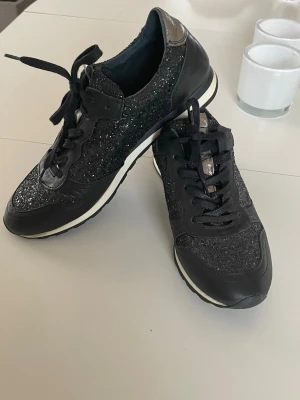 Svarta glittriga sneakers 41 - Snygga svarta sneakers med glittriga detaljer och svarta skosnören. Skorna har en låg profil och vit sula med svart kant. Gulddetaljer. Stl 41. Märke Cobler. Fint skick. 