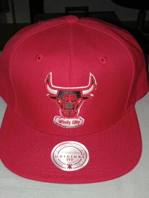 Röd Chicago Bulls snapback keps - Snygg röd snapback keps från Mitchell & Ness med Chicago Bulls-logga broderad framtill och texten 'Windy City' under. Klassisk platt skärm och justerbar passform. Perfekt för dig som gillar streetwear och sportig stil.