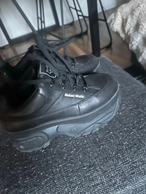 Svarta Naked Wolfe sneakers - Chunky svarta sneakers från Naked Wolfe med hög platåsula och logga på sidan samt bak på hälen. Skorna är tillverkade i svart läder med svarta skosnören och har en futuristisk vibe. Perfekta för dig som gillar statement-skor och streetwear.