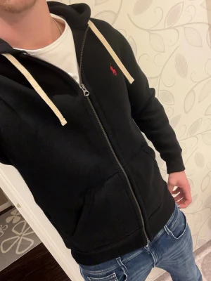 Svart hoodie från Ralph Lauren - Säljer en svart hoodie från Ralph Lauren med dragkedja och röd logga på bröstet. Hoodien har vita snören i huvan och två fickor framtill. Perfekt för en avslappnad och stilren look. Helt oanvänd hör av er om frågor