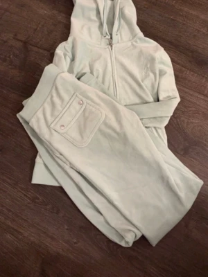 Ljusgrönt velourset Juicy Couture - Supermysigt ljusgrönt loungewear-set från Juicy Couture i mjuk velour. Setet består av en hoodie med dragkedja och matchande byxor med ficka bak. Perfekt för chill dagar hemma eller när du vill vara extra comfy. Den är som ny då jag inte har kunnat använda den för den är lite för stor för mig 