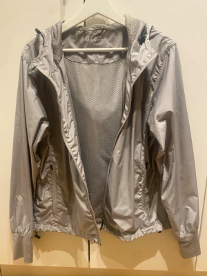 Ljusgrå vindjacka från Triberg - Ljusgrå vindjacka från Triberg med huva och meshfoder. Jackan har dragkedja framtill, två sidofickor med dragkedja och justerbar nederkant. Perfekt för blåsiga dagar och enkel att packa ner i väskan. Stilsäker och funktionell design.