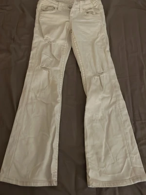 Beiga utsvängda jeans med slitningar - Snygga vita jeans med utsvängda ben och små hål. Jeansen har låg midja och subtila slitningar vid knäna för en trendig look. Perfekta för dig som gillar stockholmstil och vill ha till tillexempel sommaren ett par ljusare färg på jeansen. Barn stolek ifrån Gina!