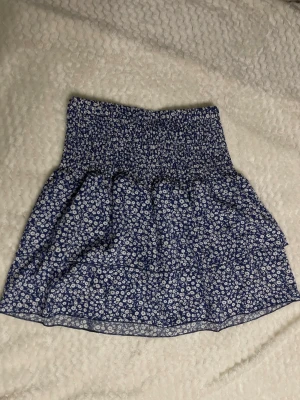 Blå blommig kortkjol med volang - Supersöt blå kortkjol med små vita blommor och bred smock i midjan. Kjolen har volangdetaljer och är perfekt för dig som gillar en feminin och avslappnad stil. Passar till både sneakers och sandaler.