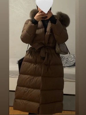 Brun lång dunjacka med pälshuva - Säljer en brun lång dunjacka med stor pälskantad huva och midjebälte. Jackan har en quiltad design och går ner till vaderna, perfekt för kalla vinterdagar. Stängs med dragkedja och knytband i midjan för extra passform.