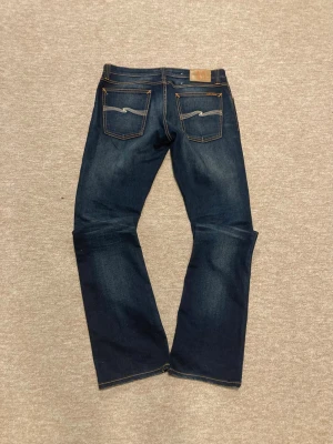 Mörkblå jeans från Nudie Jeans - Säljer ett par mörkblå jeans från Nudie Jeans med klassisk femficksmodell och snygga slitningar framtill. Jeansen har raka ben och kontrastsömmar samt Nudie-logga på bakfickorna. Tillverkade i bomull för en skön känsla.