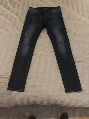 Mörkblå slim fit jeans från Jack & Jones - Snygga mörkblå slim fit jeans från Jack & Jones med klassisk femficksdesign och subtila slitningar på framsidan. Jeansen har en skinnpatch bak i midjan och är tillverkade i stretchigt denimtyg för extra komfort.