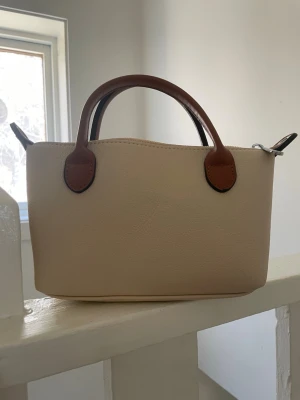 Beige handväska från meet me there - Stilren beige handväska med bruna& handtag och avtagbar axelrem. Väskan har en klassisk, rektangulär form och är tillverkad i ett slätt, läderliknande material. Perfekt för dig som gillar enkel och tidlös design. Mått:höjd 13cm, längd 20 cm