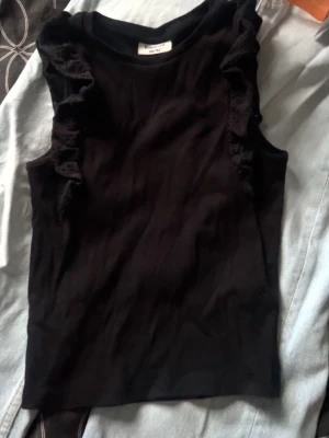 Svart ärmlös topp med volang - Snygg svart ärmlös topp från Kappahl med dekorativa volanger längs axlarna. Tillverkad i mjuk bomull som känns skön mot huden. Perfekt att styla med jeans eller kjol för en trendig look.