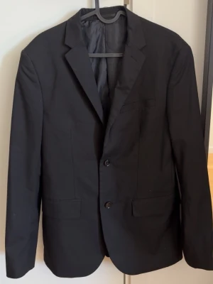 Klassisk svart kavaj med två knappar - Snygg svart kavaj med två knappar framtill och klassisk krage. Kavajen har två fickor med lock och en bröstficka. Tunn fodring på insidan och stilren passform som passar till många olika looks. Normal slim fit passform 