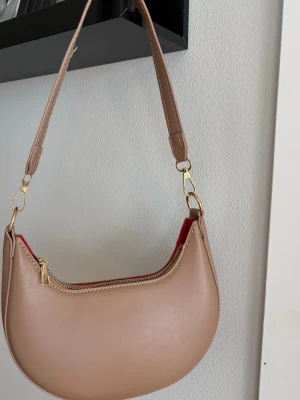 Beige/ ljusbrun axelremsväska  - Stilren beige/ ljusbrun handväska i halvmåneform med avtagbar axelrem och guldfärgade metalldetaljer. Väskan har dragkedja upptill och röd insida som ger en snygg kontrast. 