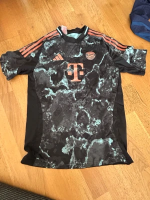 Bayern München Adidas matchtröja - Cool Bayern München matchtröja från Adidas med svart och turkosmönstrad design och orange detaljer. Tröjan har korta ärmar, rund hals och klassiska Adidas-ränder på axlarna. Klubbmärke och sponsorlogga på bröstet. Perfekt för fotbollsträning eller support.