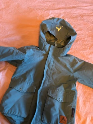 Blå vindjacka från McKinley - Snygg blå vindjacka från McKinley med huva och stora fickor framtill. Jackan har tryckknappar och dragkedja, samt en stilren logga på ena fickan. Perfekt för blåsiga dagar och outdoor-aktiviteter.