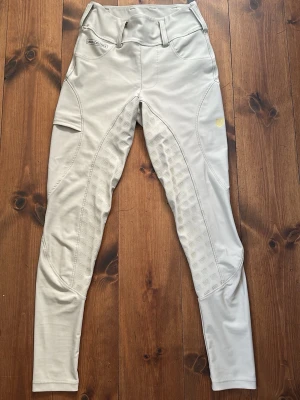 Beige ridleggings från Maya Delorez - Beiga ridleggings från Maya Delorez i modellen ”performance”. Ridbyxorna har en medel hög midja, bälteshällor, fullskoning i silikon och praktisk mobilficka på vänstra låret. De har bara använts några fåtal gånger och är därför i väldigt fint skick. ((Säljer även ett annat par liknande i marinblå, storlek xs))