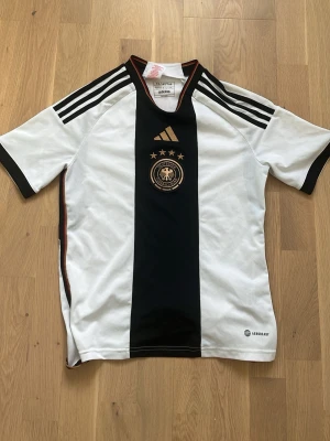 Tysklands fotbollströja Adidas - Säljer en vit och svart Tyskland fotbollströja från Adidas med tre stjärnor och förbundsmärket på bröstet. Tröjan har svarta ränder på axlarna, korta ärmar och en bred svart mittpanel. Tillverkad i lätt och ventilerande material.