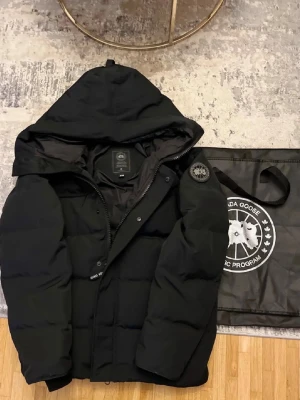 Svart pufferjacka från Canada Goose - Säljer en svart pufferjacka från Canada Goose med huva och klassisk patch på ärmen. Jackan har en stilren design med breda kanaler och dragkedja framtill. Perfekt för kalla dagar när du vill hålla värmen och se snygg ut.