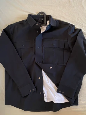 Svart overshirt från sail racing - Stilren svart overshirt från saik racing med klassisk krage, tryckknappar och två stora bröstfickor med lock. Overshirten är tillverkad i ett slätt och tåligt material som ger en clean look. Perfekt att bära över en t-shirt för en avslappnad men snygg stil.