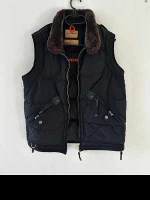 Svart dunväst Parajumpers med fuskpäls - Svart dunväst från Parajumpers med bruna detaljer och mjuk fuskpäls runt kragen. Västen har två snedställda fickor med dragkedja och tryckknapp, samt robust dragkedja framtill. Perfekt för lager-på-lager och streetstyle. Priset kan justeras självklart!!