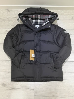 Svart pufferjacka från Burberry - Svart pufferjacka från Burberry med avtagbara ärmar och huva. Jackan har rutigt foder i grått, vitt och rött samt dragkedja och tryckknappar framtill. Två snedställda fickor och Burberry-logga på ärmen. Perfekt för kalla vinterdagar.