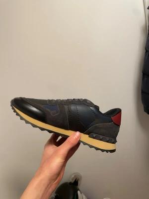 Valentino Garavani sneakers svart/grå - Snygga sneakers från Valentino Garavani i svart och mörkgrå mocka och läder, med detaljer i marinblått och en röd hälflik. Yttersulan är beige med svarta nitar runt hälen och har en räfflad undersida för extra grepp. Perfekta för dig som gillar streetwear och exklusiva märken.
