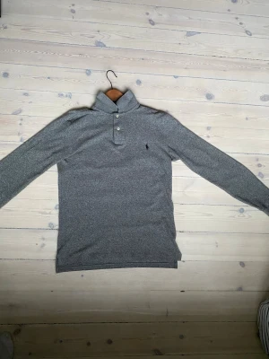 Grå långärmad pike från Polo Ralph Lauren - Snygg grå långärmad pike från Polo Ralph Lauren i slim fit. Skjortan har klassisk krage, två knappar vid halsen och den ikoniska broderade loggan på bröstet. Tveka inte och göra av dig