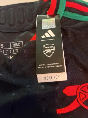 Arsenal svart matchtröja Adidas S - Svart Arsenal matchtröja från Adidas med gröna och röda detaljer vid kragen, röd kanonlogga och vit Emirates Fly Better-tryck på bröstet. Slim fit-modell med HEAT.RDY-material som andas. Perfekt för fotbollsträning eller supporterliv.
