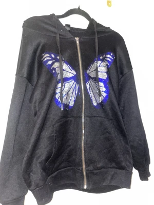 Svart hoodie med fjärilstryck - Cool svart hoodie med dragkedja och stor glittrig fjäril i blått, vitt och silver på bröstet. Huva med snören och känguruficka framtill. Perfekt för dig som vill sticka ut med en unik detalj.