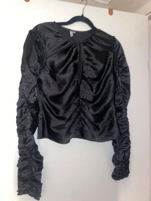 Svart satinlik topp med rynkade ärmar - Trendig svart topp i glansigt satinliknande material med långa, rynkade ärmar och rynkade detaljer framtill. Croppad modell med rund halsringning, perfekt för en cool och stilren look.