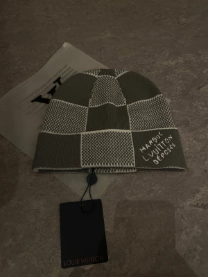 Gråvit mössa - Säljer en exklusiv gråvit mössa med rutigt mönster och broderad text på uppviket. Mössan är tillverkad i en lyxig mix av 49% kashmir, 40% ull och 11% silke. Perfekt accessoar för dig som vill ha något unikt och stilrent.