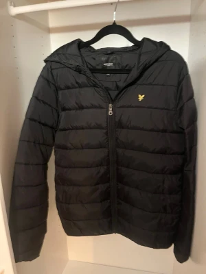 Svart pufferjacka från Lyle & Scott - Svart pufferjacka från Lyle & Scott med huva och dragkedja framtill. Jackan har ett quiltat mönster och den klassiska gula loggan på bröstet. Perfekt för kalla dagar och enkel att matcha med olika outfits.