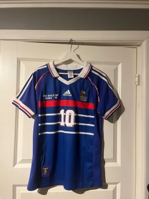 Frankrike Zidane VM 98 tröja Adidas - Klassisk blå fotbollströja från Adidas, Frankrikes landslag, VM 1998. Nummer 10 och Zidane på ryggen, broderad tupp-logga och vita/röda detaljer. Kortärmad med V-ringning och tryckta ränder över bröstet. Perfekt för fotbollsfans!