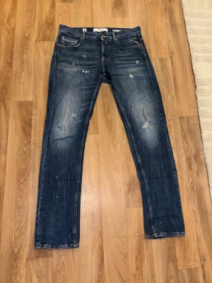 Replay Jeans W29 L32 - Säljer ett par blå jeans från Replay med snyggt slitna detaljer och klassisk femficksmodell. Jeansen har raka ben och en cool tvätt med ljusare partier på låren och knäna. Perfekta för en avslappnad och trendig look. Modellen på sista bilden är 181cm och väger 79kg.