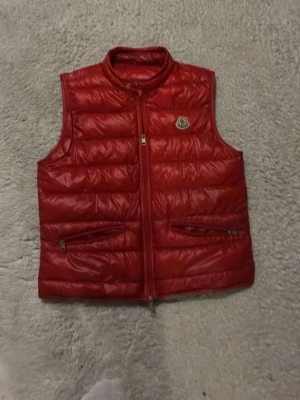 Röd dunväst från Moncler - Snygg röd dunväst från Moncler med quiltad design och dragkedja framtill. Västen har två dragkedjefickor och Moncler-logga på bröstet. Perfekt för lager-på-lager och ger en cool vibe till din outfit. och har ett hål kolla sista bilden