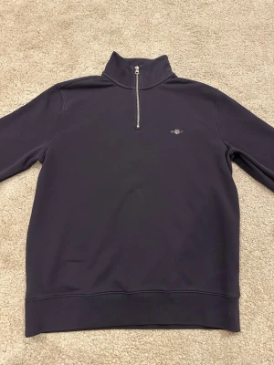 Mörkblå halfzip sweatshirt från GANT - Snygg mörkblå sweatshirt från GANT med halv dragkedja och hög krage. Liten broderad logga på bröstet och ribbade muddar. Perfekt för en stilren och avslappnad look. Mjukt material på insidan för extra komfort.