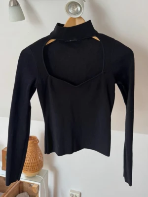 Svart långärmad topp med öppen byst - Trendig svart långärmad topp med hög hals och snygg öppen detalj över brösten.  Toppen har en croppad passform och är perfekt för att skapa en cool och stilren look. Materialet känns mjukt och stretchigt, vilket gör den bekväm att bära hela dagen.