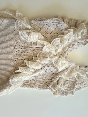 Beige stickad linnetopp med spets - Supersöt beige stickad linnetopp med vackra spetsvolanger längs axelbanden och halsringningen. Toppen har ett detaljerat mönster i stickningen och är ärmlös, perfekt för en romantisk och trendig look.