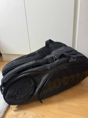 Wilson svart ryggsäck för tennis - Säljer en svart ryggsäck från Wilson, perfekt för tennisutrustning. Väskan har flera fack, inklusive isolerade innerfack för racketar och tillbehör. Tillverkad i slitstarkt syntetmaterial med justerbara axelremmar och diskret logga på sidan.