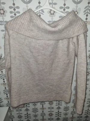 Beige stickad tröja med bred krage - Mysig beige stickad tröja med bred, nedvikt krage och långa ärmar. Tröjan har en mjuk och fluffig känsla, perfekt för kyliga dagar. Passar dig som gillar en avslappnad men ändå stilren look.