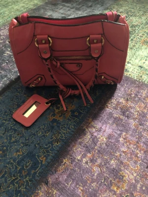 Rosa m handväska i konstläder - Cool rosa handväska mjukt läder med flätade handtag, guldfärgade metalldetaljer och dragkedja framtill. Väskan har en avtagbar tag och klassisk city-modell vibe. Perfekt för dig som vill sticka ut med en statementväska.