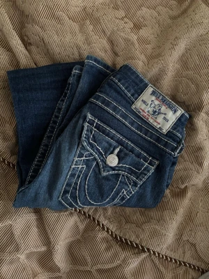 True Religion bootcut jeans blå - Säljer ett par blå bootcut jeans från True Religion med kontrastsömmar och snygga fickdetaljer bak. Jeansen är lågmidjade. 