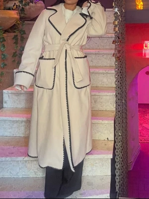 Off white/ Beige kappa med svarta detaljer - Stilren kappa med svarta detaljer. Kappan har knytskärp i midjan och är så elegant. Köpte i frankrike, säljer då jag känner att den är lite för lång på mig som är 155cm. 