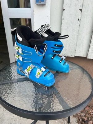 Blåa pjäxor från Lange - Säljer ett par blåa pjäxor från Lange med svarta och vita detaljer. De har fyra spännen i metall och grönt för extra justering samt en svart/vit rem upptill med tydlig Lange-logga. Insidan är vadderad för komfort och passform. Perfekta för skidåkning i vinter.