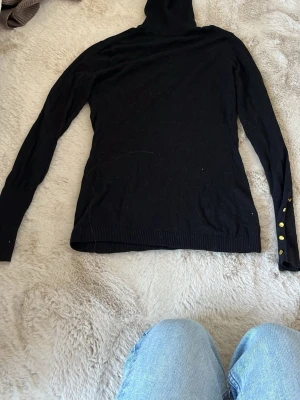 Svart polotröja med guldknappar - Snygg svart stickad polotröja med långa ärmar och ribbade muddar. Ärmsluten är dekorerade med fyra guldiga knappar som ger en extra touch. Perfekt till jeans eller kjol för en stilren look. har så många polotröjor och behöver sälja nån och de blev tyvärr denna! älskar verkligen den o perfekt särskilt med guld smycken!